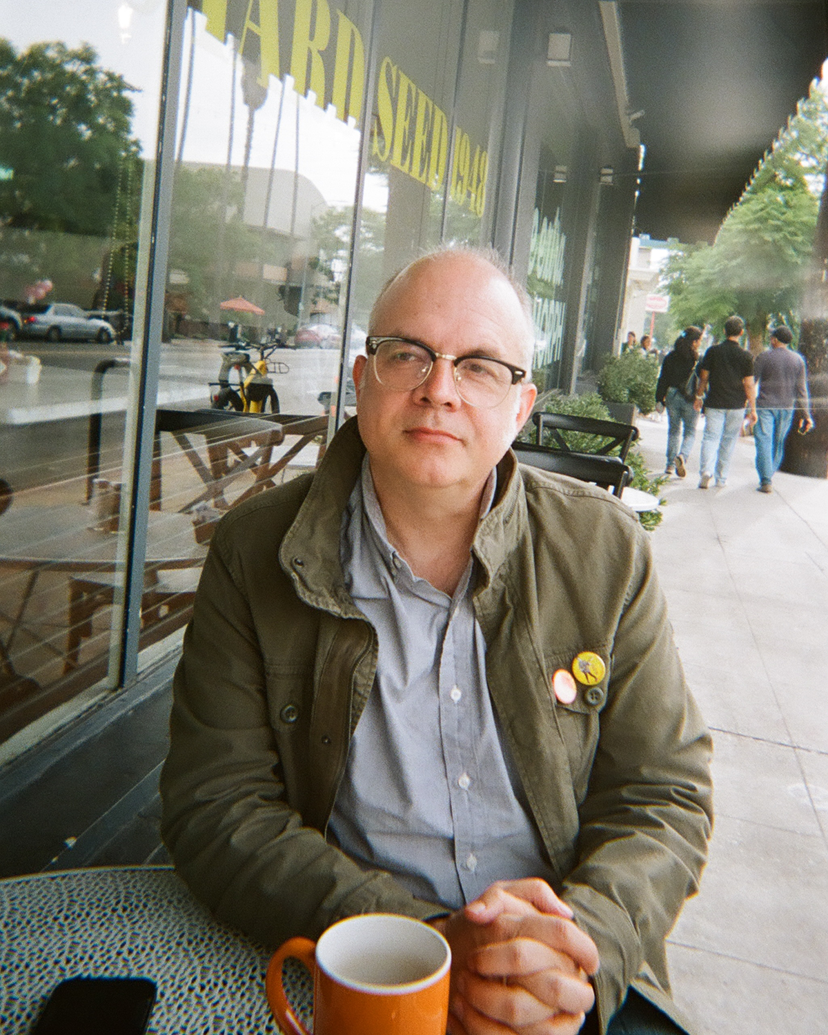 Hines sits with hands clasped at a small outdoor cafe table. He stares off into the distance thoughtfully. He has an orange mug in front of him. He wears green jacket with bright buttons on the right side over a sensible bluish-gray button-up.