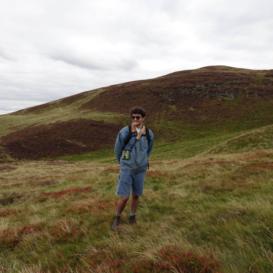 Daniel Heumann stands in a field in front of a rolling hill in rainy Edinburgh, Scotland.