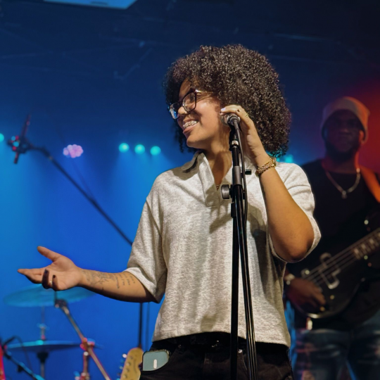 Lyric Vargas in Lisbon, Portugal. They are on stage in front of a mic, smiling with hand out.