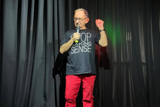 Brian Flesher holds a microphone on stage against a black curtain.