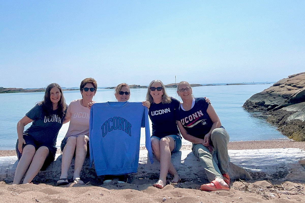 Five friends lean to the middle of the photo. They sit on a log on a beach in front of blue sky and water.