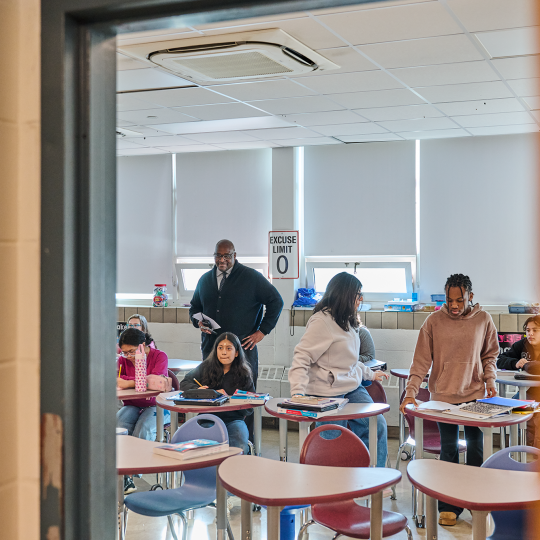 Lewis smile pleasantly in the middle of a classroom where students are taking notes.