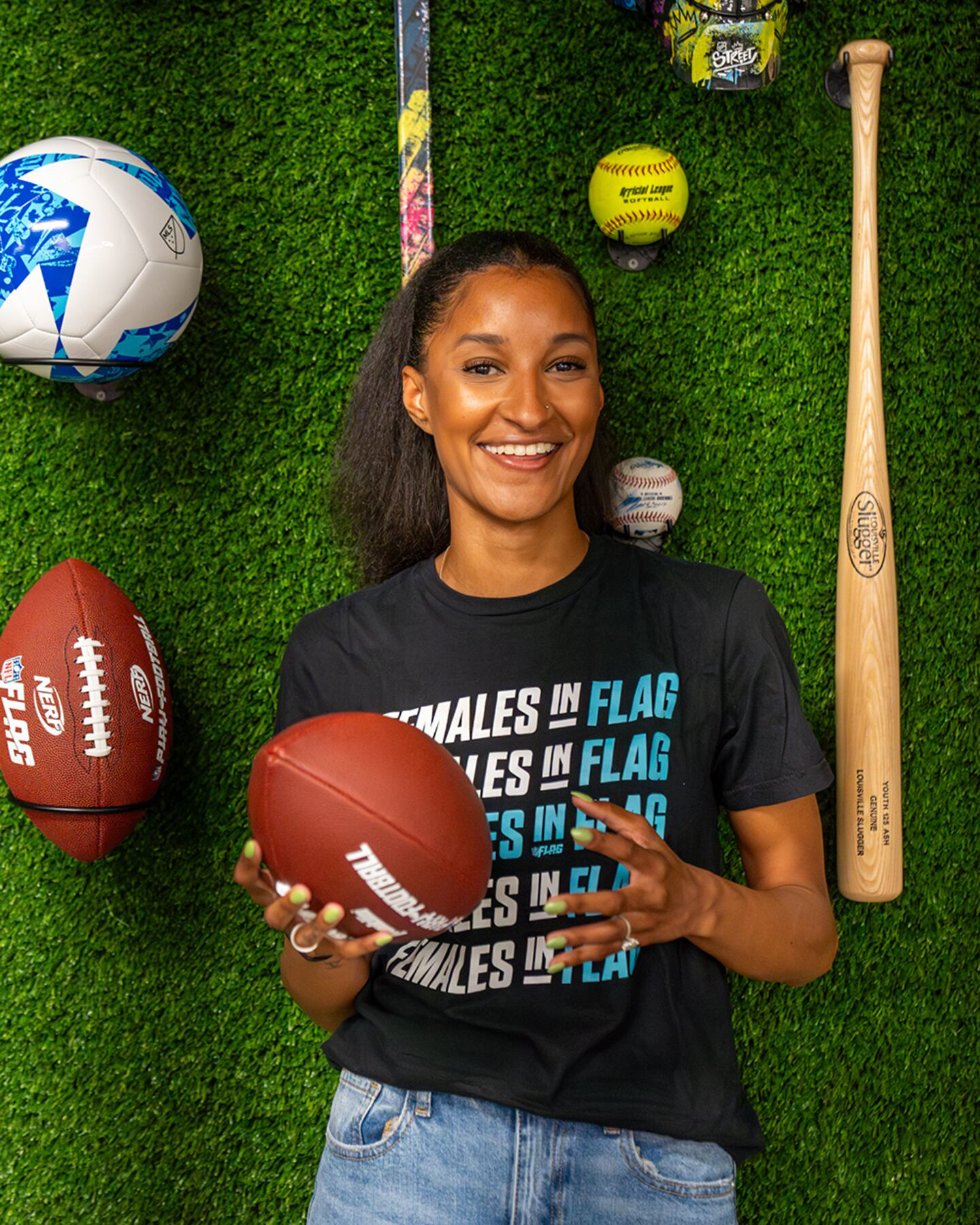 Jade Strawberry smiles and shifts a football in her hands. She wears a shirt that reads "Females in Flag". She is posed in front of a wall of green turf with baseball, baseball bat, soccer ball, football, and other sports gear.
