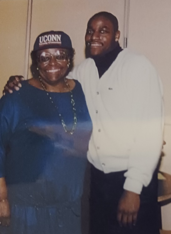 Dave Lewis with mom, Beverly, after Lewis announced his decision to attend UConn in 1989. Lewis and Beverly smile for the camera. Lewis has his arm around his mother's shoulder. Beverly wears a UConn hat and blue dress.