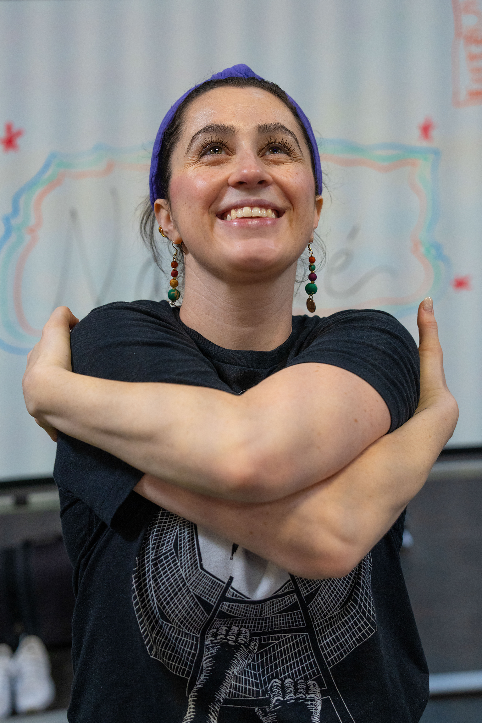 A yoga teacher smiles brightly as she looks upwards, practicing a yoga move that looks like a self-hug.