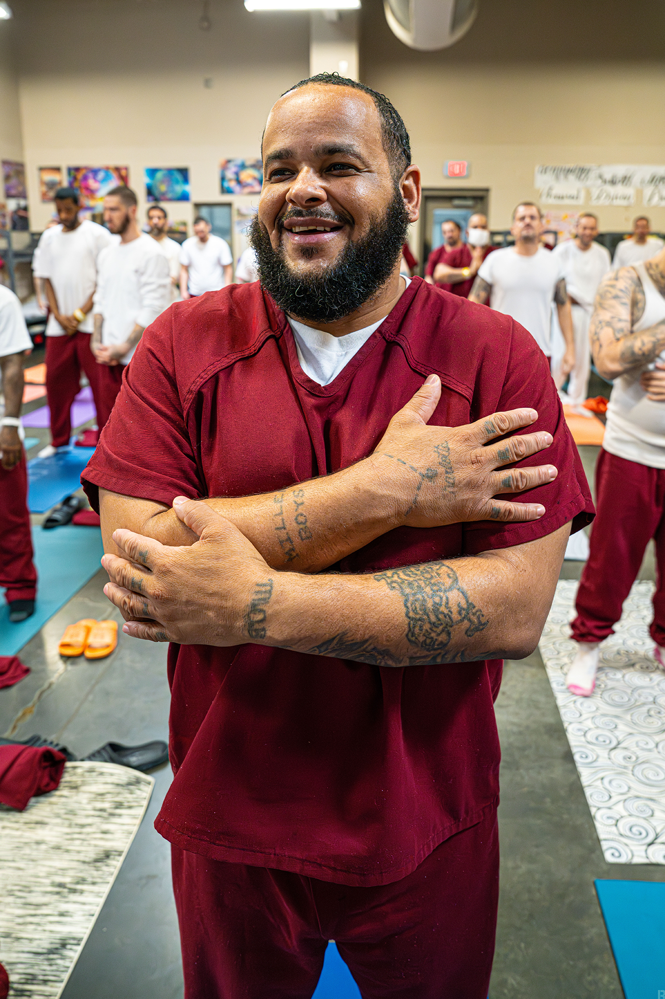 A tatooed man in a red prison jumpsuit smiles cheerily atop his yoga mat, he seems to do a yoga pose that looks like a self-hug. His fellow yogis stand on their yoga mats behind him in their yoga class.