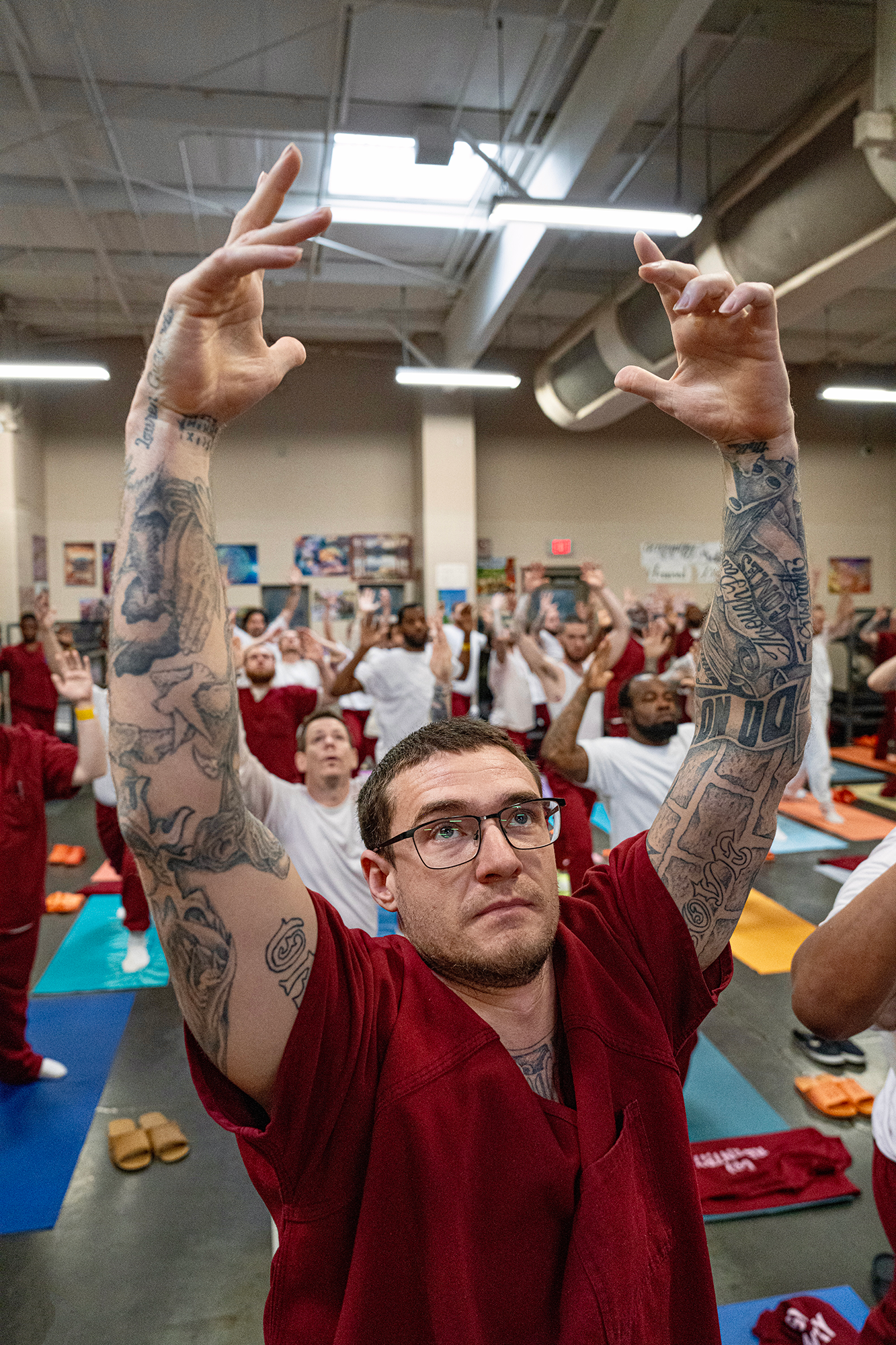A man in a red prison jumpsuit raises his tatooed arms above his head in a yoga pose. Behind him, his fellow yogis are also practicing the same pose.