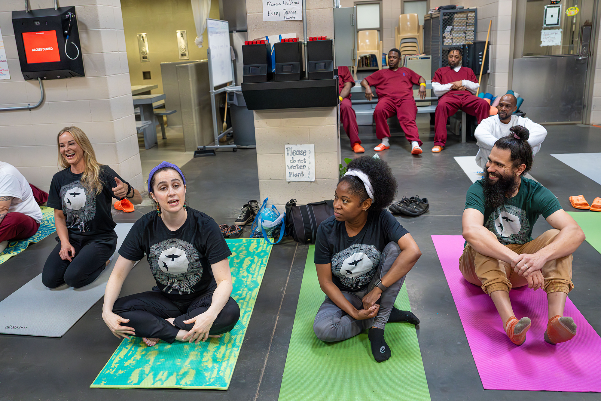A group of yoga teachers sits atop their yoga mats, looking cheery as one of them addresses the yoga class, off screen. Behind them, inmates in red jumpsuits recline in chairs in another room, just slightly out of focus.