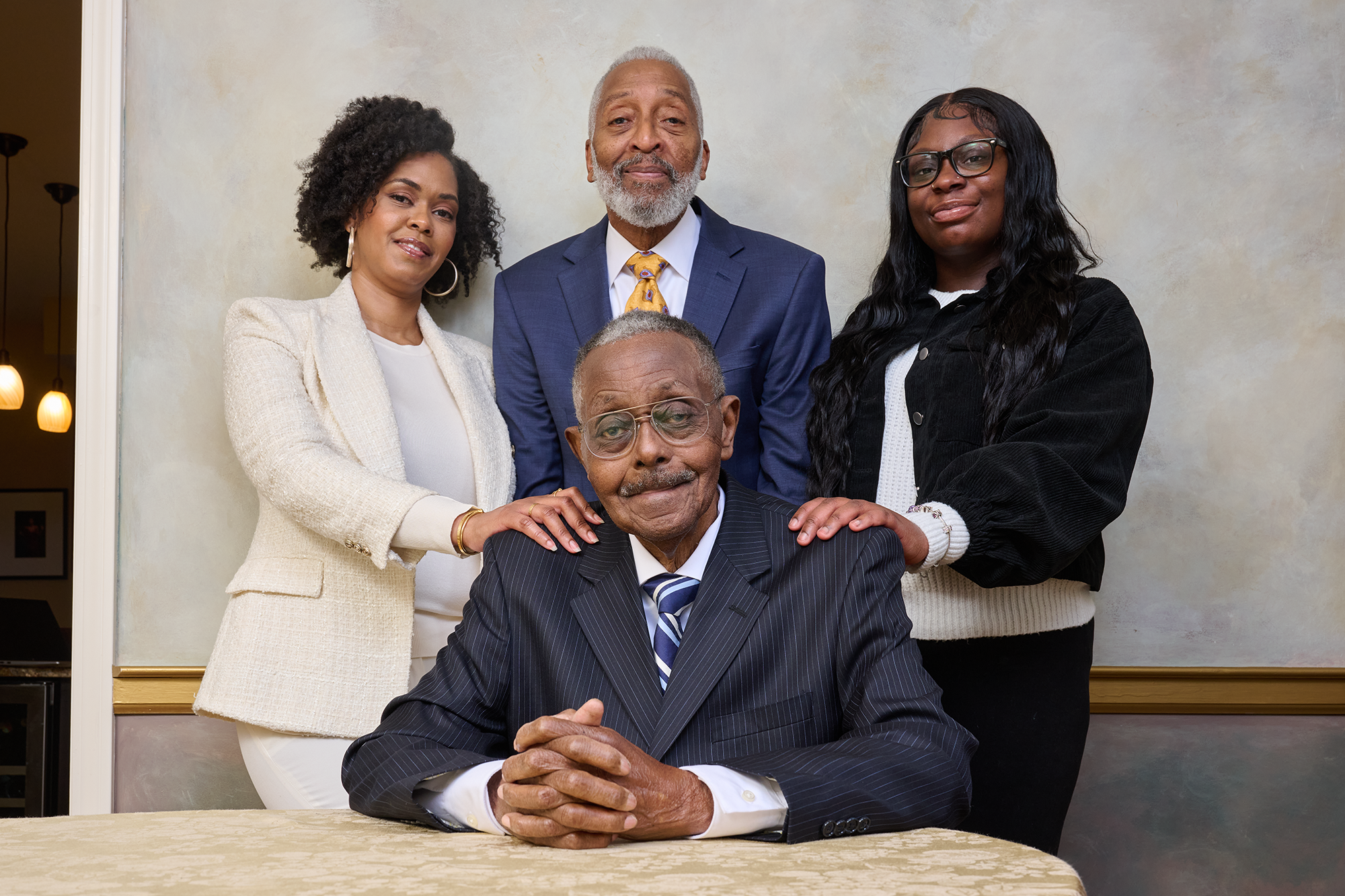 A group of four people, two men, Leonard Blanks and Otis Wade, and two women Courtney and Solé Blanks, pose proudly for a photo. They are all dressed in suits. The two women have one hand on each shoulder of the elder gentleman, Otis, sitting at a table in the foreground of the group shot.