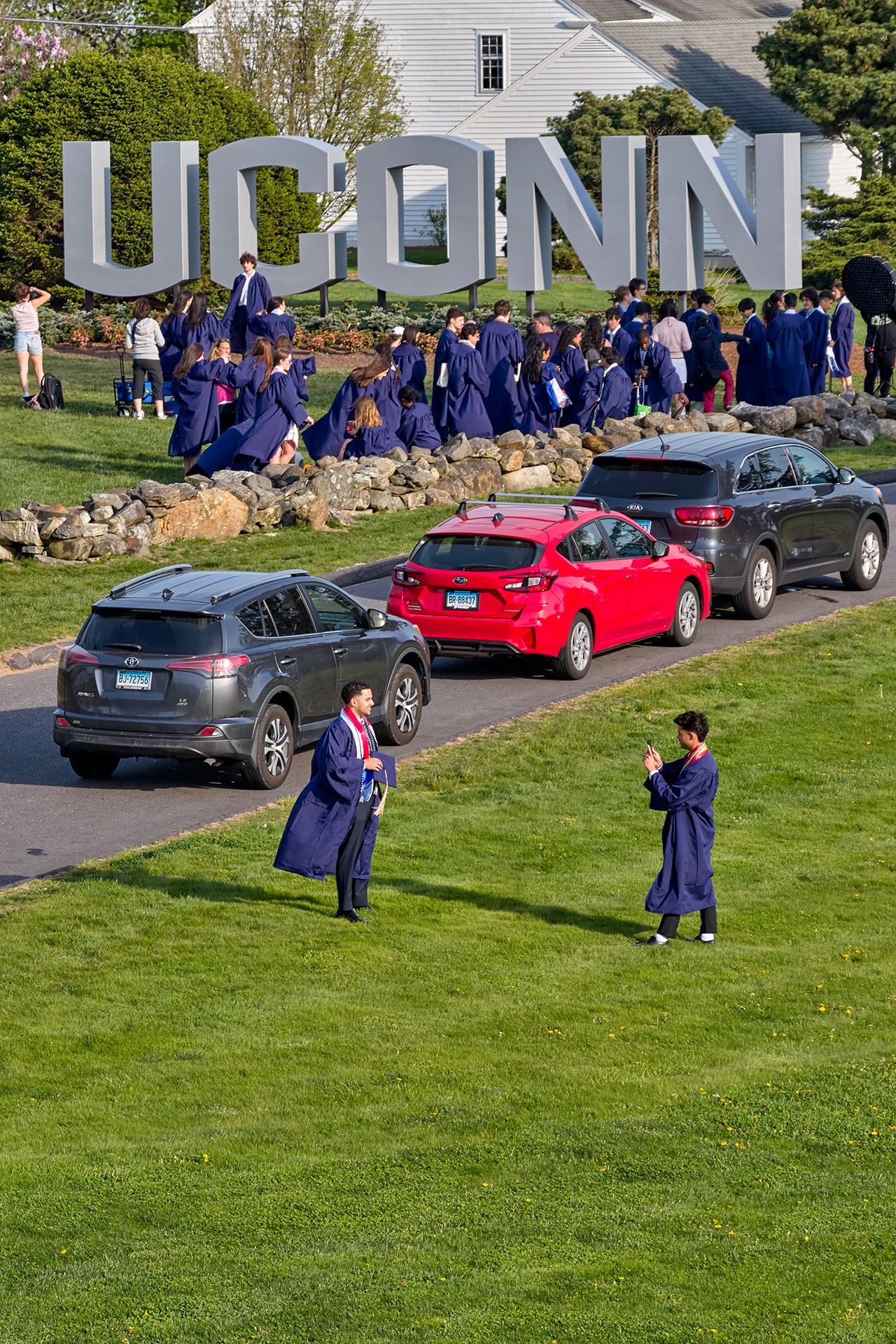 Graduates are lined up to take photos at the UConn Entryway Sign. Across the road, a graduate takes a picture of another graduate in the open grass area.