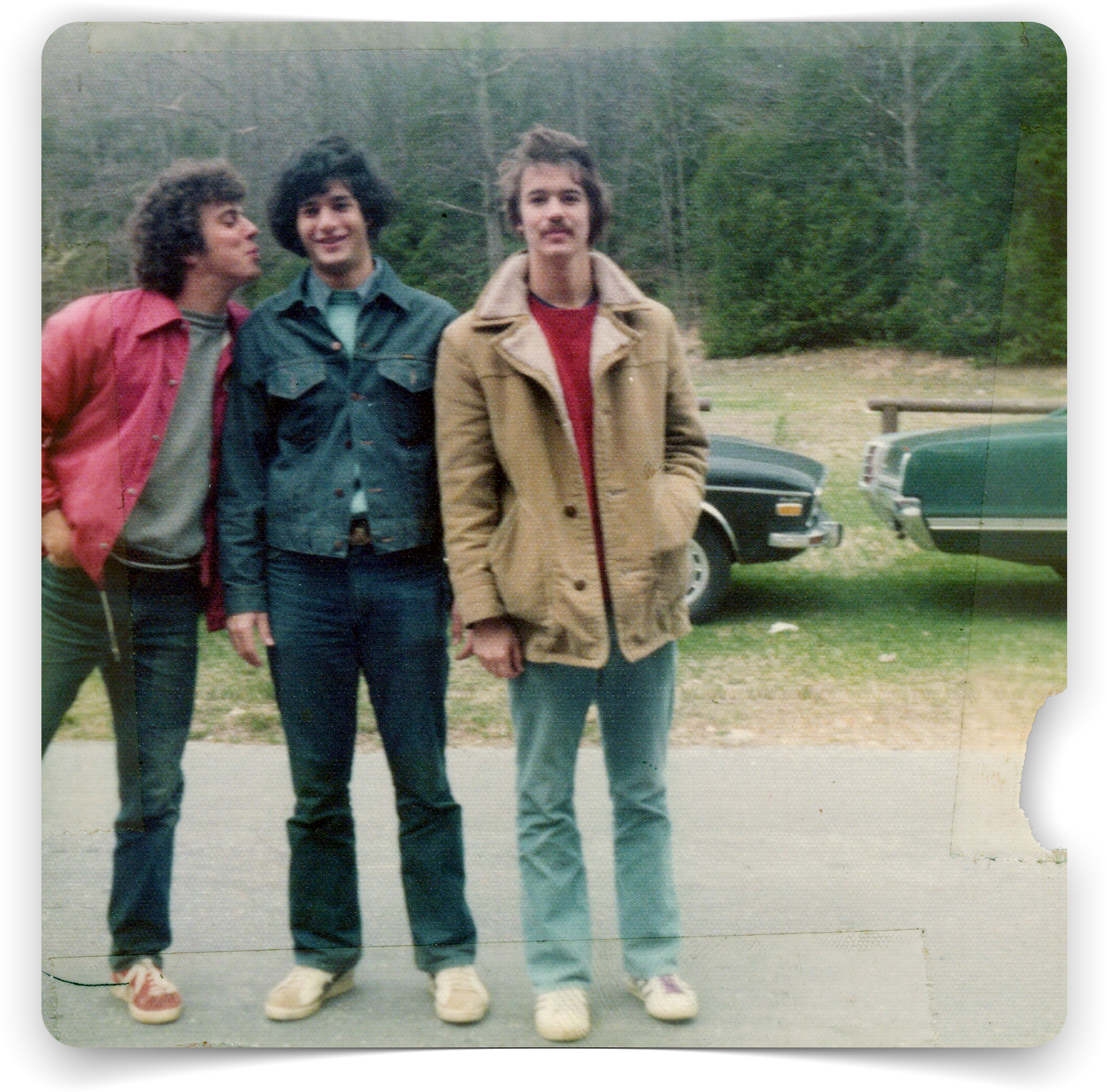 A vintage photo of three men standing together, posing playfully in front of parked cars at a campsite.