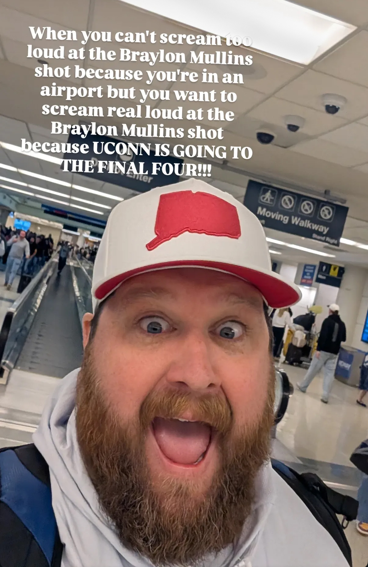 Geary in a selfie for a social media post celebrating the basketball game. He expresses a silent scream of excitement. He is at an airport and wears a hat decorated with the Connecticut state.