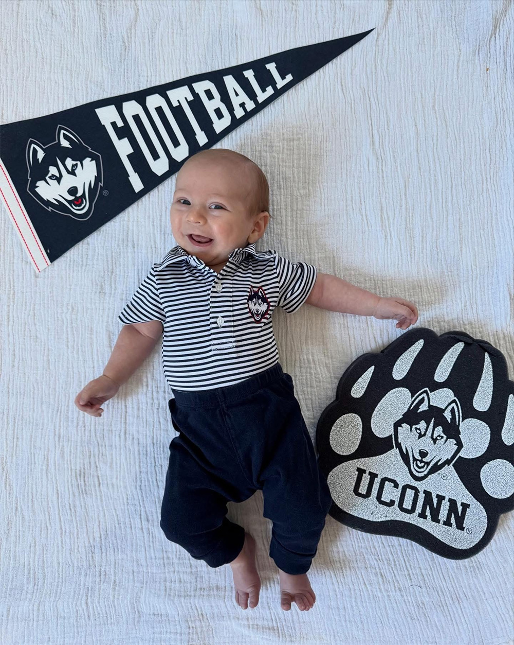 A baby in UConn Husky shirt, displayed between UConn football flag and UConn husky paw.