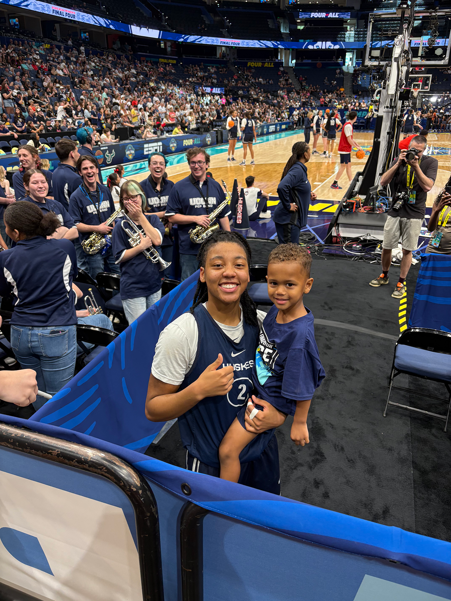 KK Arnold holds up her nephew, Talen, for a photo at a UConn game. She wears a blue vest and white shirt.