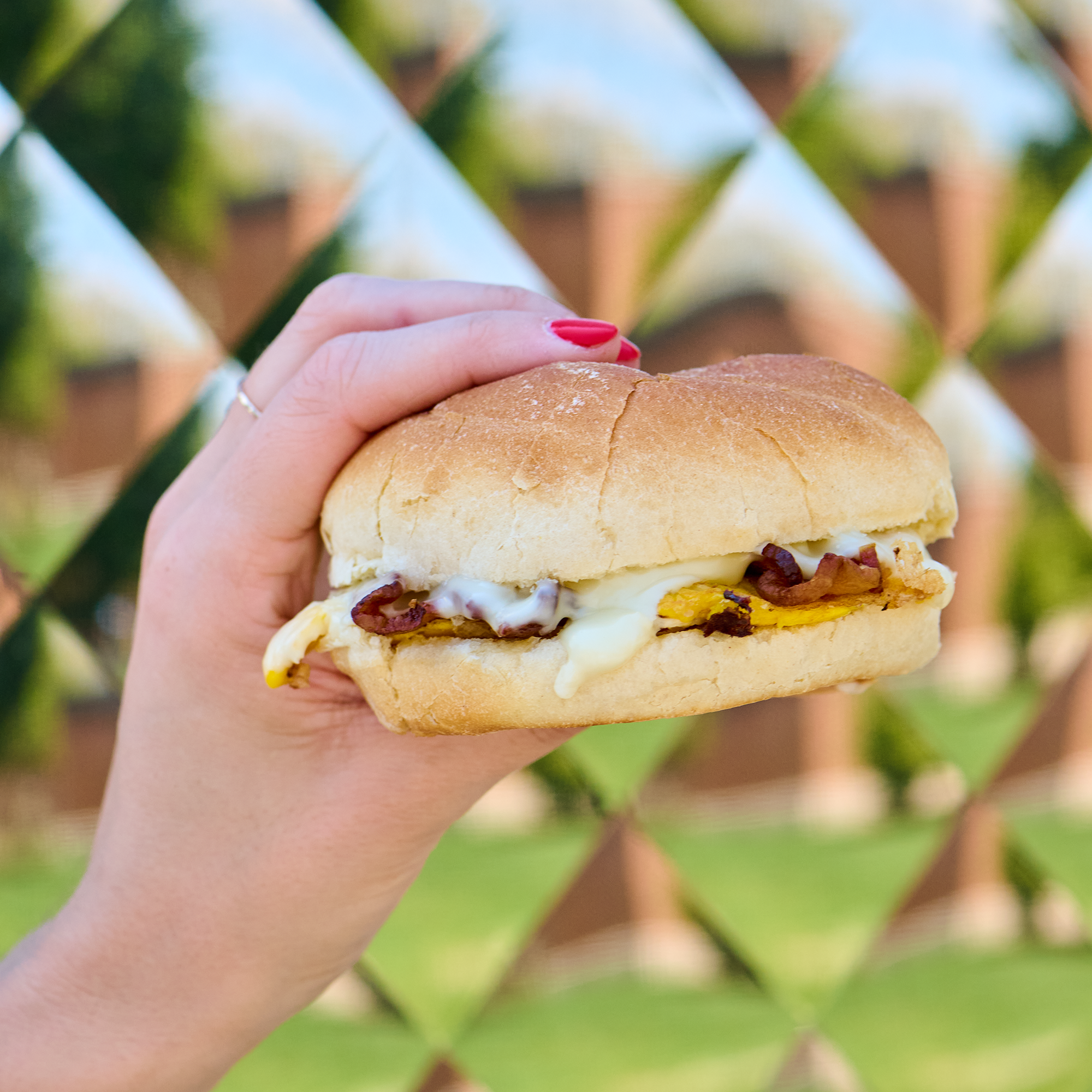 a close up shot of a sandwich with bacon, egg, cheese, and mushroom. The sandwich is held up by a hand with nails painted red. The backdrop is blurry and tiled like a kaleidoscope.
