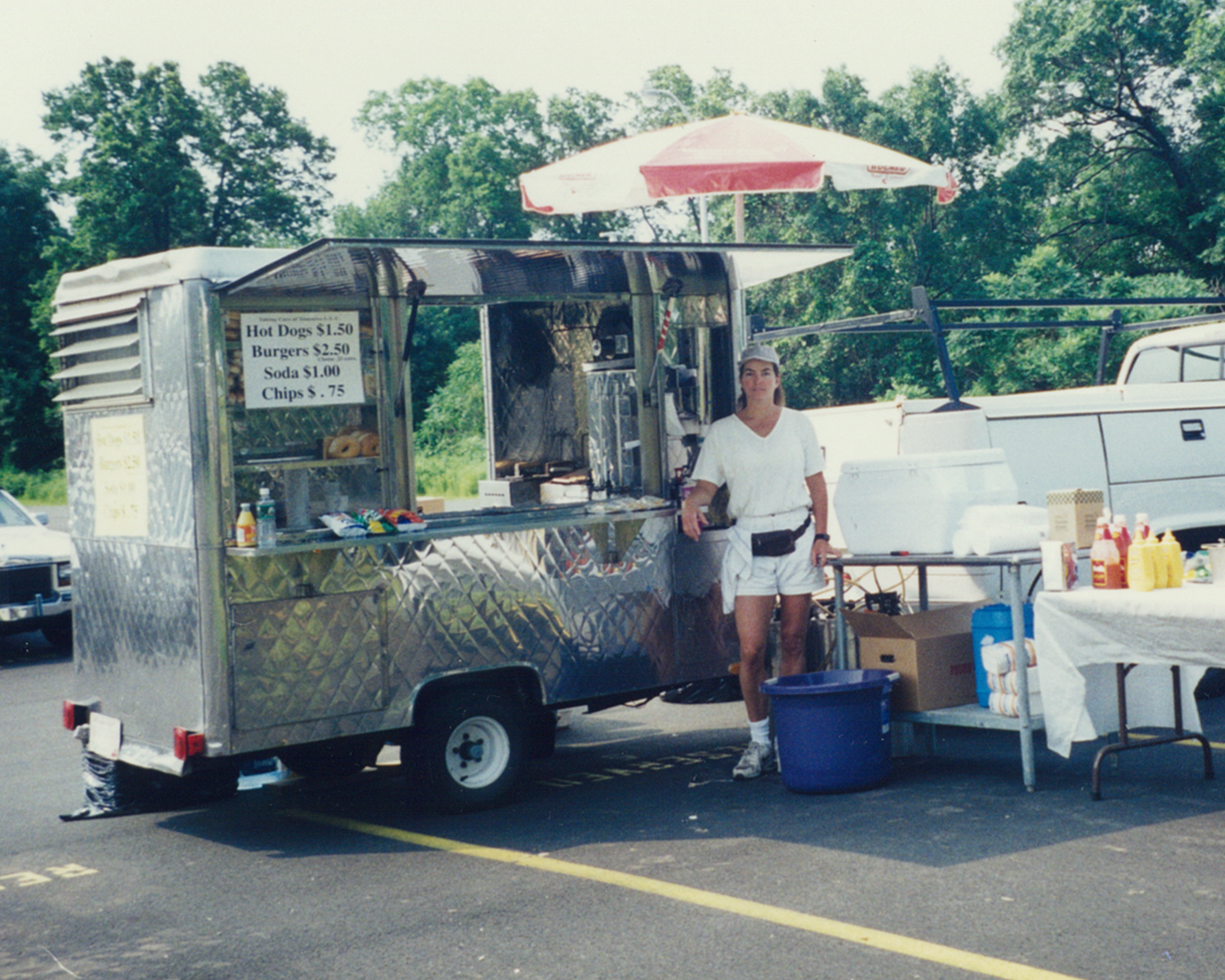 A vintage photo of a woman dressed in white with black phanny pack, standing next to a hot dog cart in a parking lot.