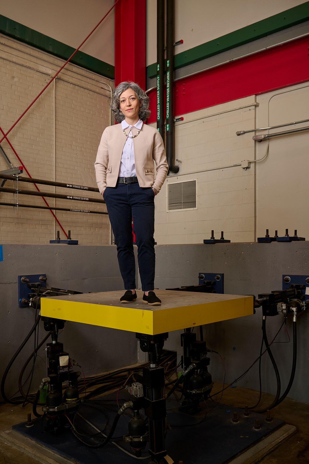 Sarira Motaref, in khaki cardigan stands on top of a yellow platform, known as a shake table. Her hands are in her pockets nonchalantly as she looks a the viewer calmly.