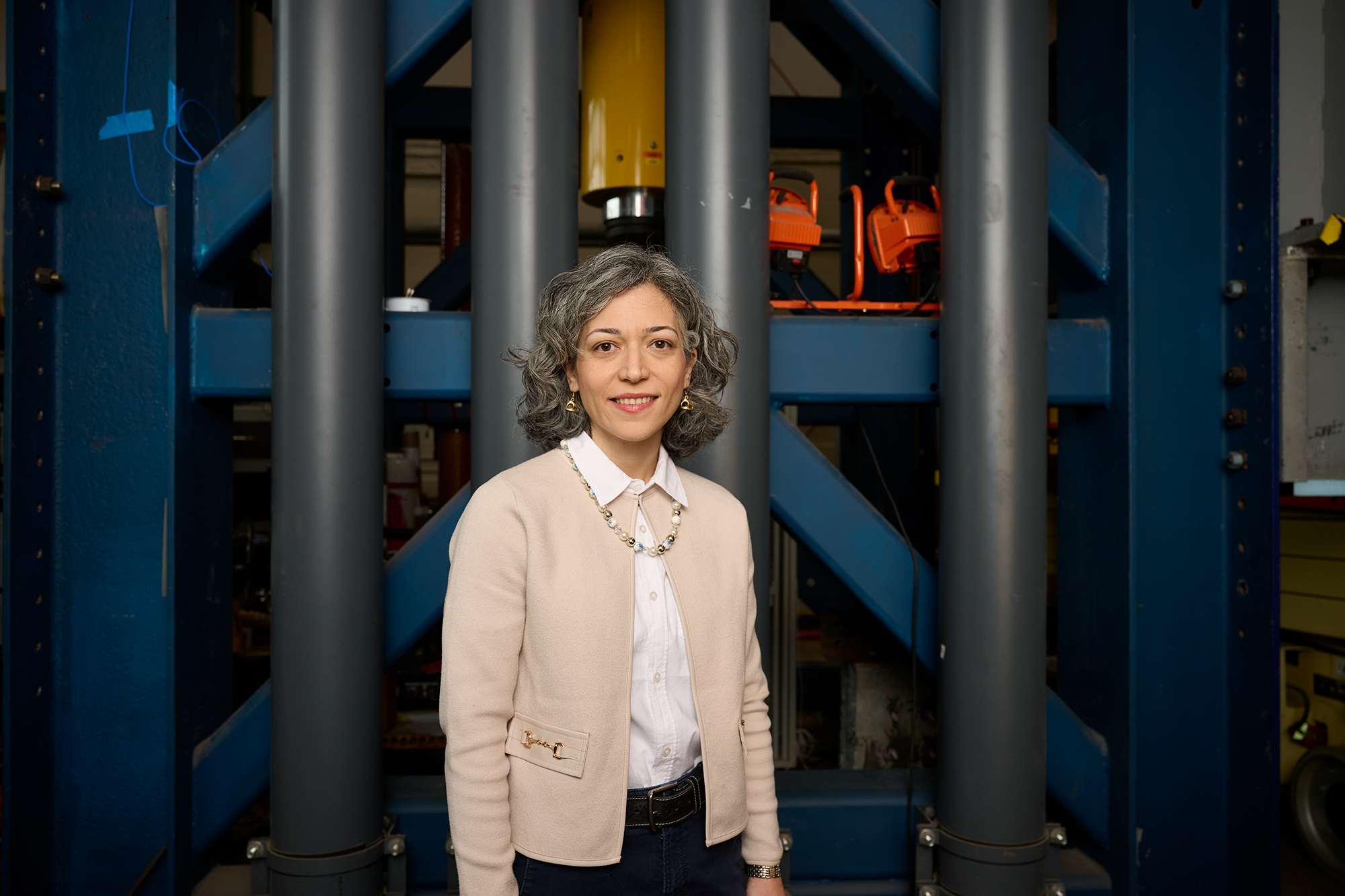 Motaref in khaki cardigan and black pants stands in front of a large metal structure.