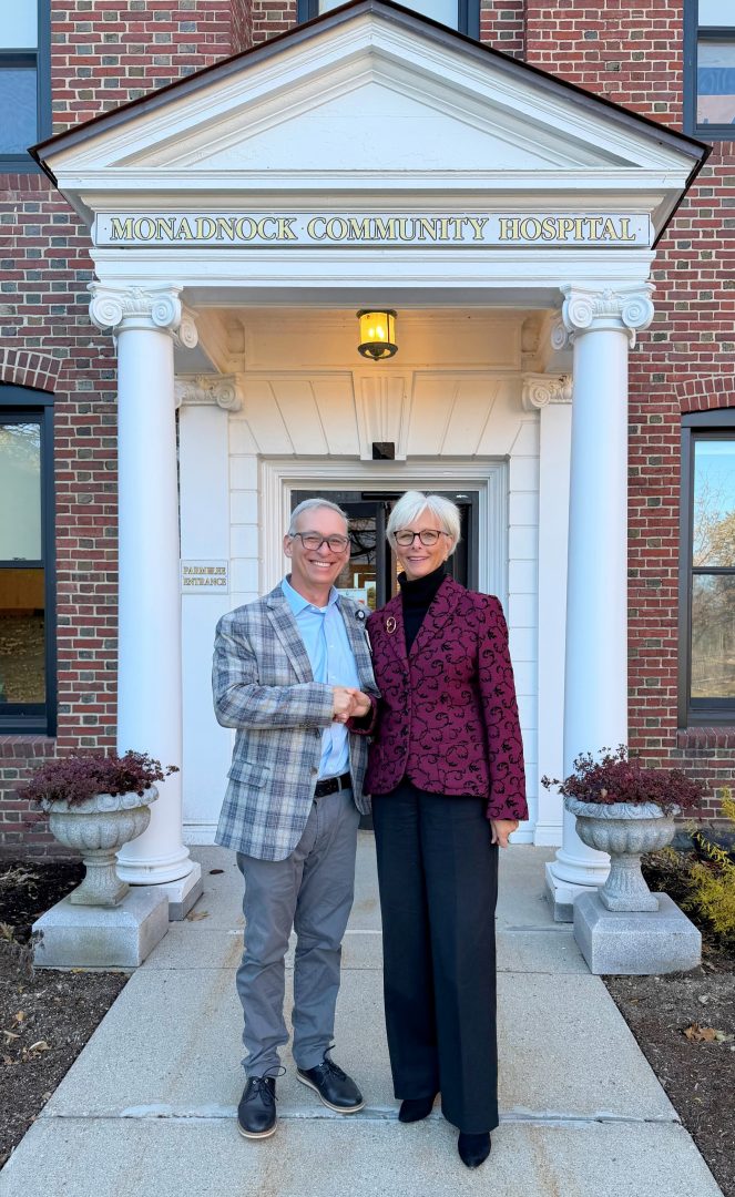 a man and woman, both with light hair and glasses wearing business attire, pose in front of Monadnock Community Hospital