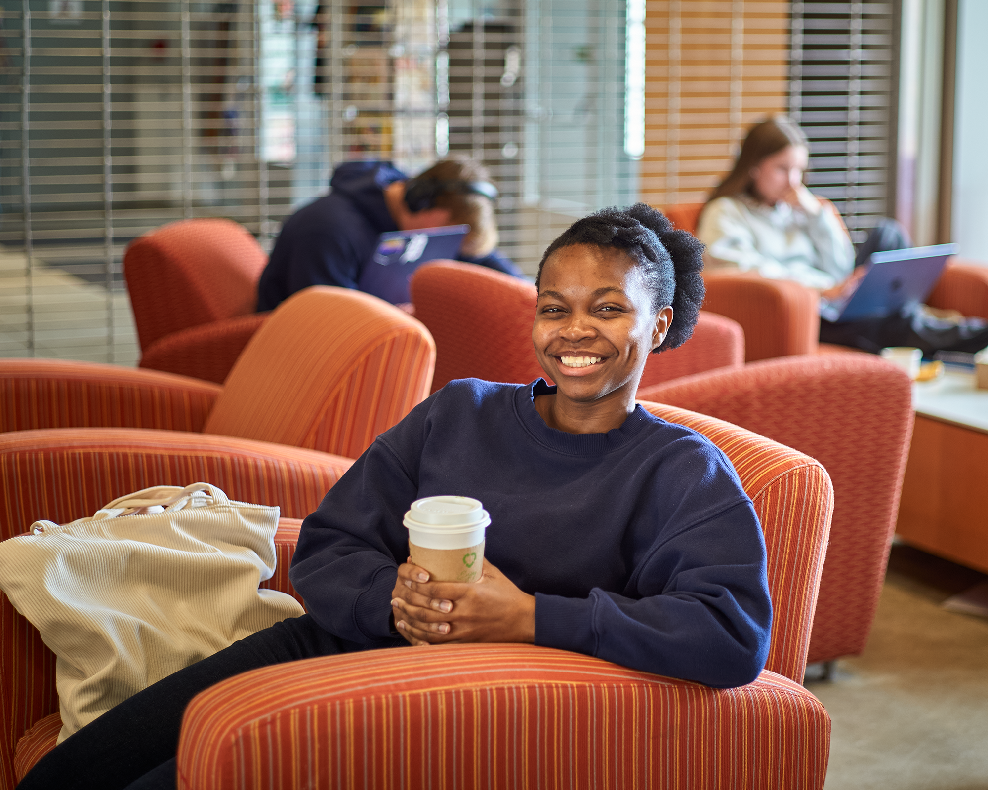 Moyo, woman in blue, sits in a soft orange armchair, holding a coffee cup. She is smiling and appears to be enjoying her time in the cozy atmosphere.