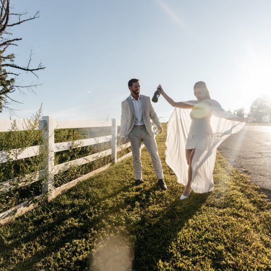 A bride and groom post in front of a white fence with bright sunlight cast over them