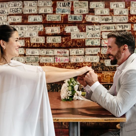 A bride and groom pose at a table in a restaurant with dollar bills adorning the wall behind them