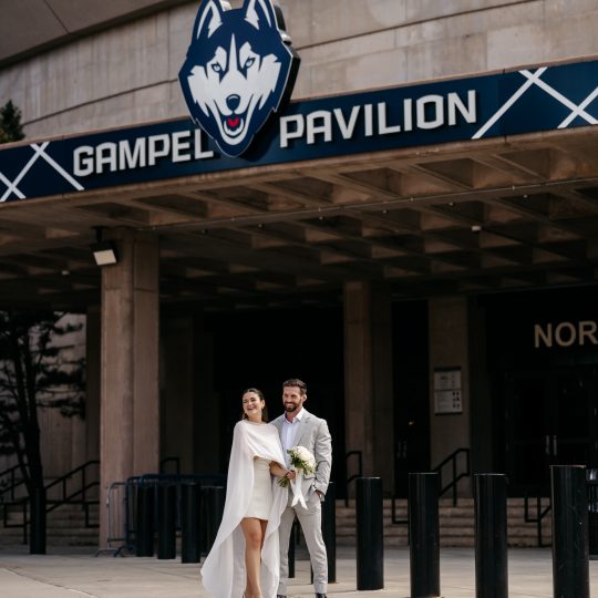 A bride and groom pose in front of Gampel Pavilion with the sign above their heads