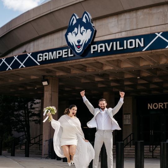 A bride and groom pose in front of Gampel Pavilion with the sign above their heads