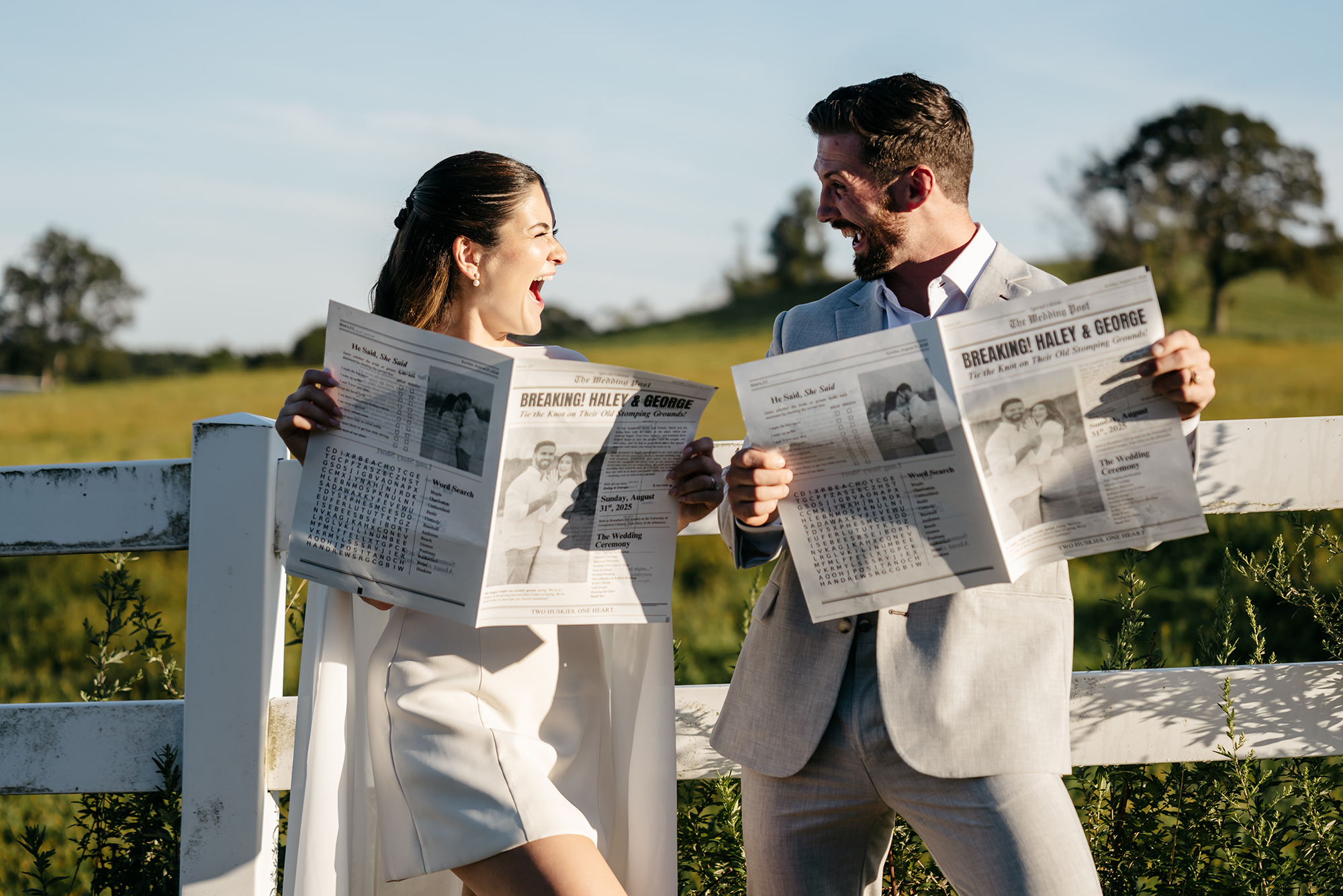 A bride and groom, Haley Stein and George Andrews, pose in front of a white fence in Horse Barn Hill on a sunny day. They pose excitedly and look at each other with energetic expressions. They are both holding custom newspaper with articles about their relationship, with big headline that read: "Breaking! Haley & George"