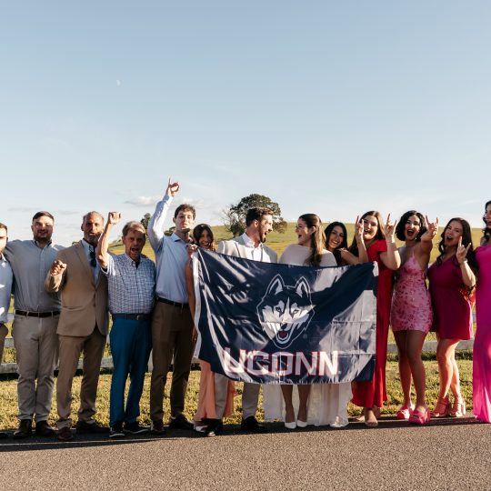 A group of UConn alums pose with a UConn flag in front of Horsebarn Hill