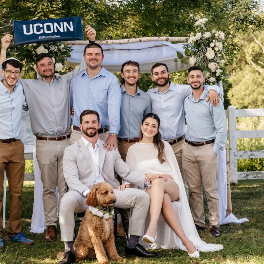 A group of wedding guests pose behind a seated groom and bride with a dog and a UConn banner