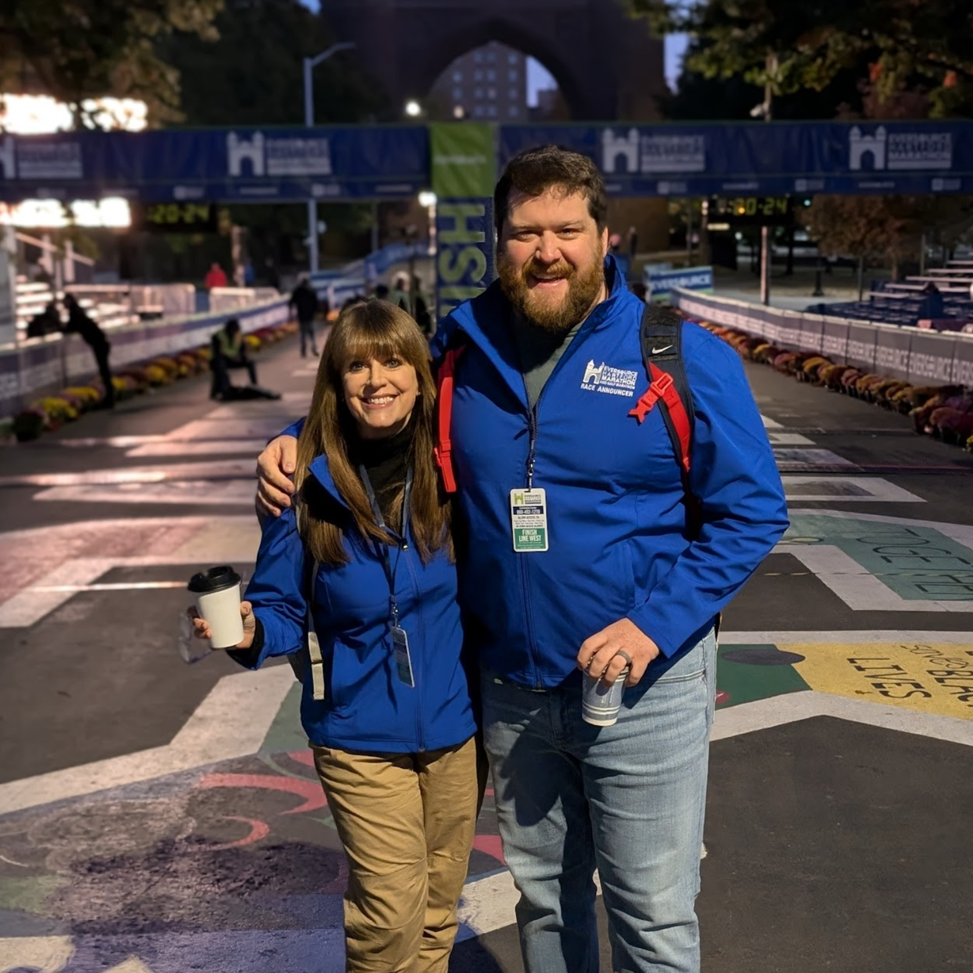 Geary in blue coat and red backpack posing with a woman holding a coffee cup.