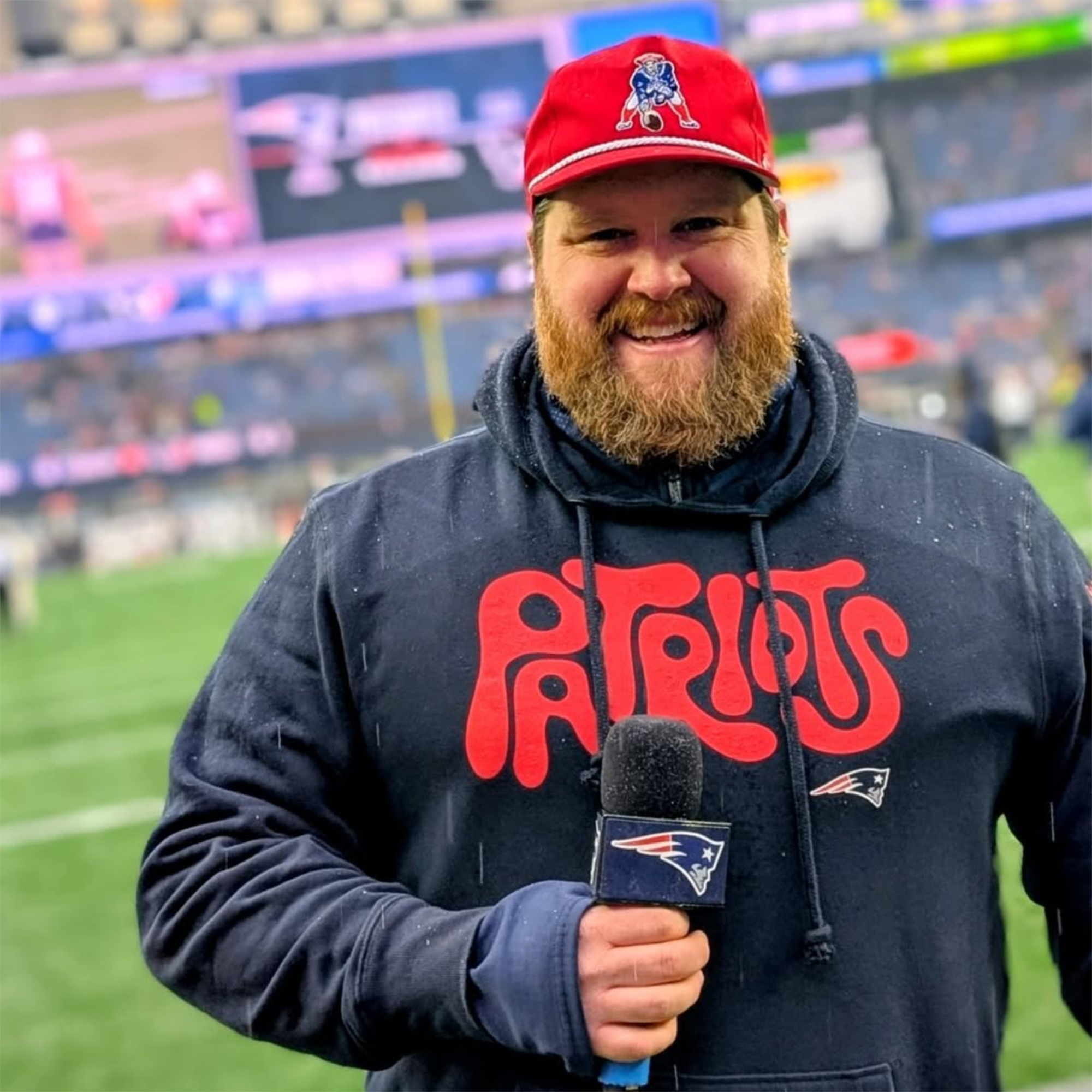 Geary in bright red cap and dark graphic hoodie with bright red Patriots signage on it. He is smiling and holding his microphone. He is in front of a football field.