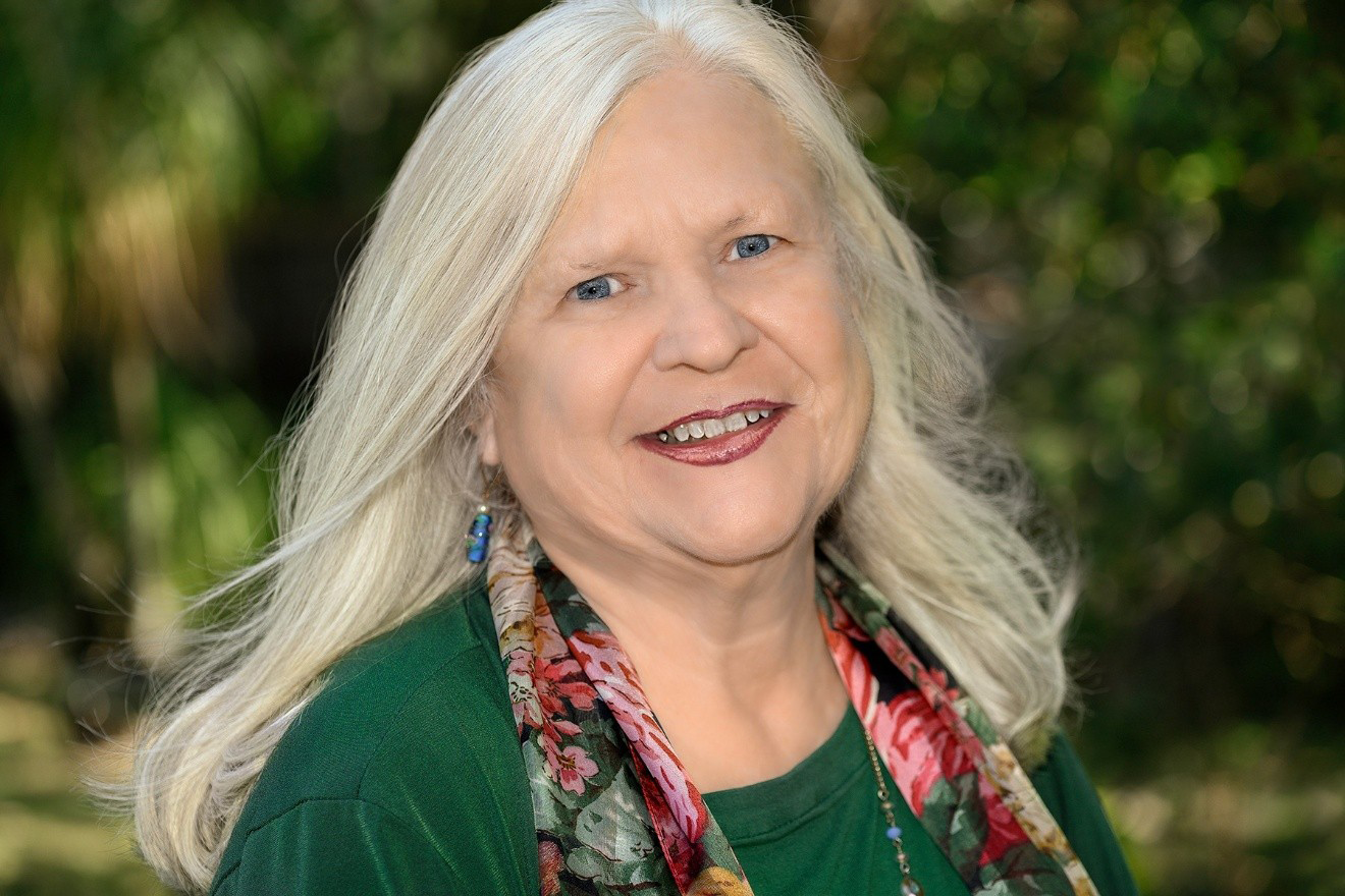 Brenda Bean smiles for the camera, she is wearing a green shirt against a nature backdrop.