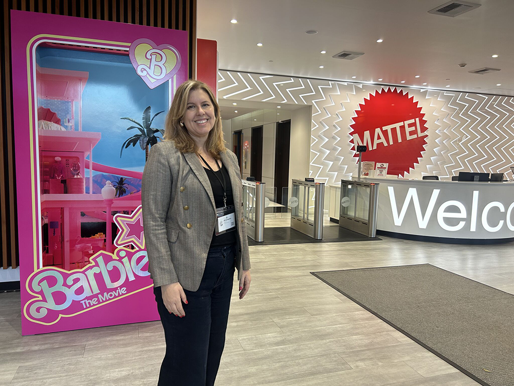 A photo of Jill Arvanitis, she is smiling for the camera in front of the Mattel logo and a Barbie box.
