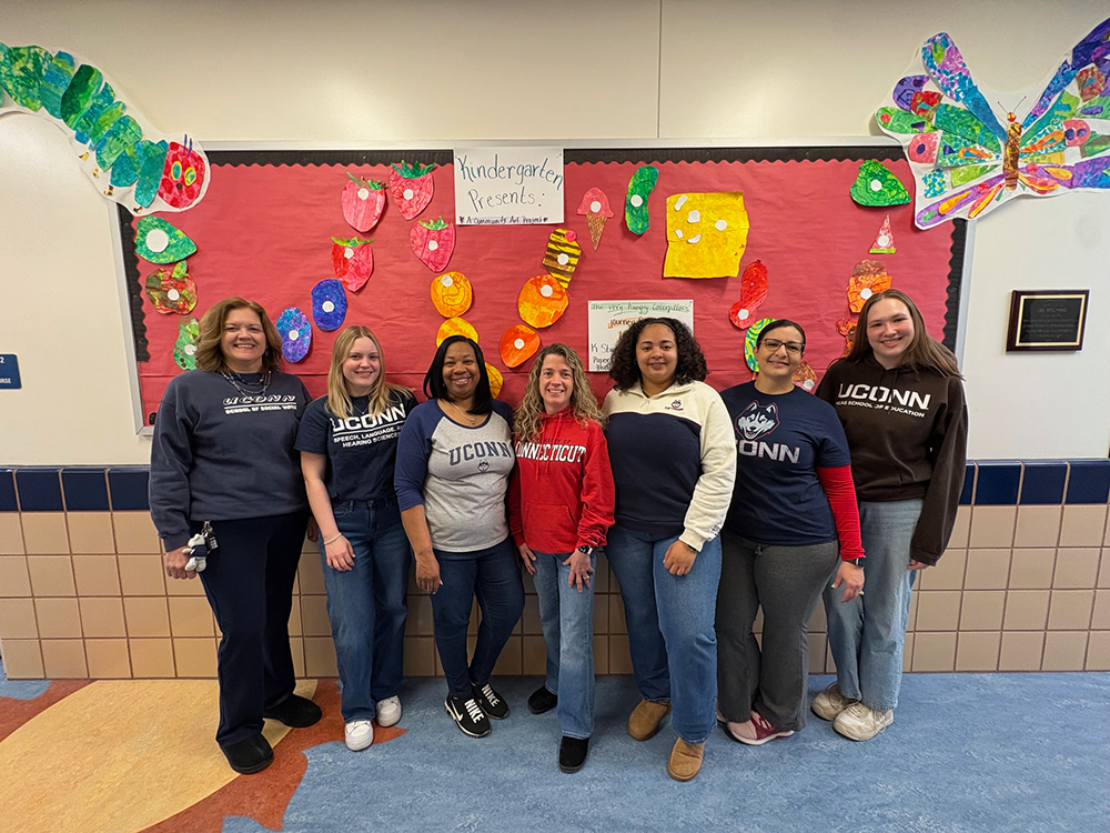 A group of UConn-trained educators pose for the photo against a brightly decorated corkboard.