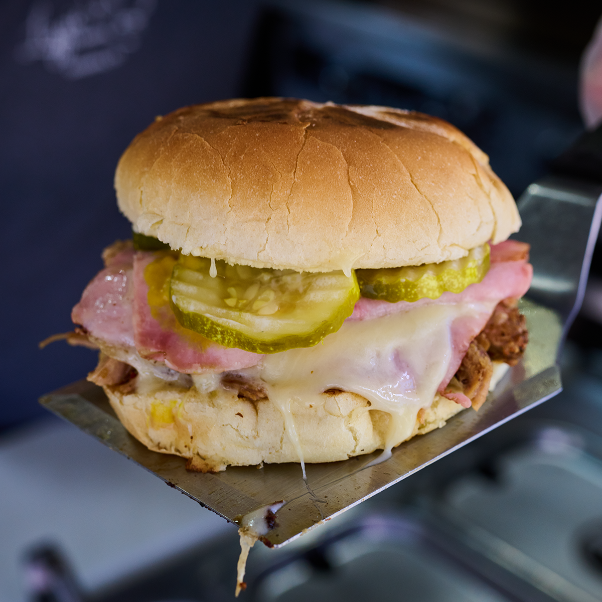 A yummy looking cuban sandwich displayed on top of a spatula. The cheese is melted and gooey, the meat is plentiful, and the pickles look juicy.