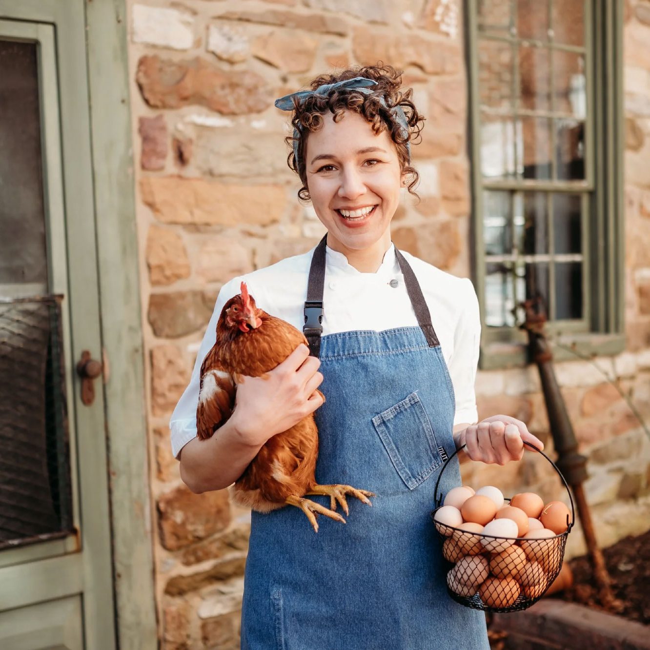 Rebecca Elman in jean-material apron smiles for the camera. She holds a chicken in one arm and a basket of eggs in the other.